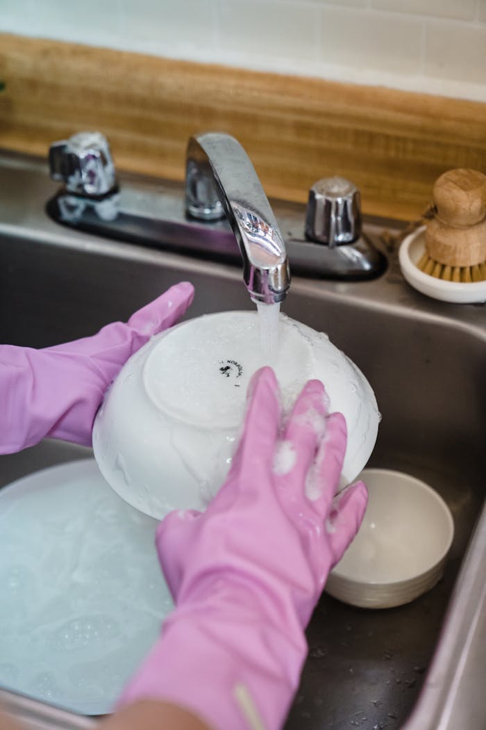 Hands wearing pink gloves washing dishes in a kitchen sink with running water.